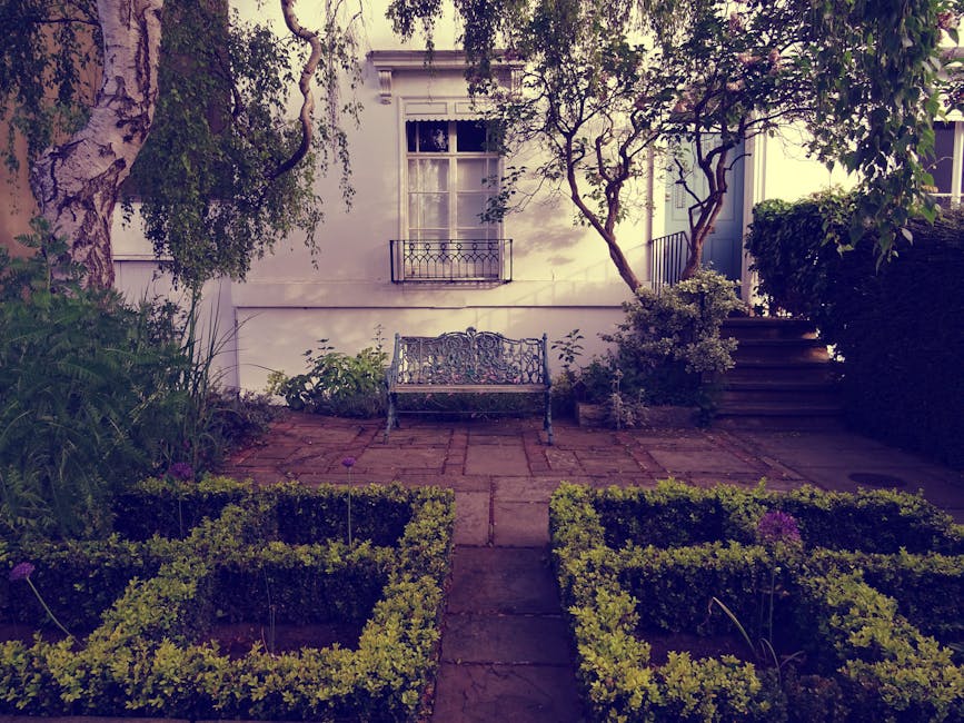 A small urban garden featuring a paved brick patio area with a decorative metal bench placed against a white wall with a window and small balcony above. The garden is bordered by neatly trimmed hedges