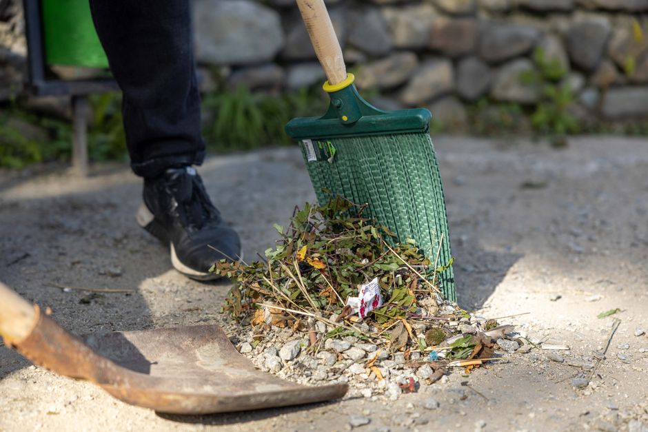 A person wearing black trousers and sneakers is using a green push broom to sweep dry leaves, twigs, and small garden debris into a pile on a gravel pathway. The pathway is bordered by a low stone wal